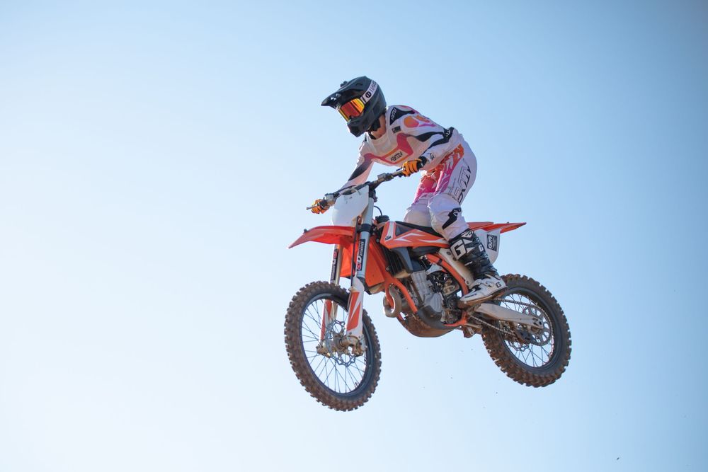 A motocross rider performs a high jump on an orange KTM dirt bike against a blue sky during competition.