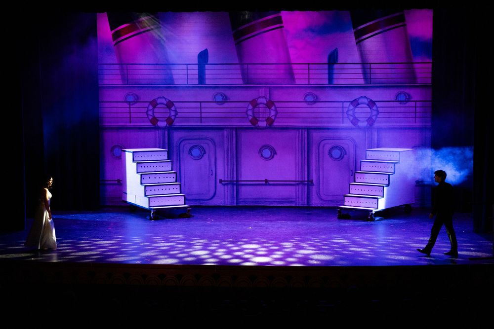 Stage lighting creates a dramatic purple and blue atmosphere during a theater production with spotlights and set pieces.