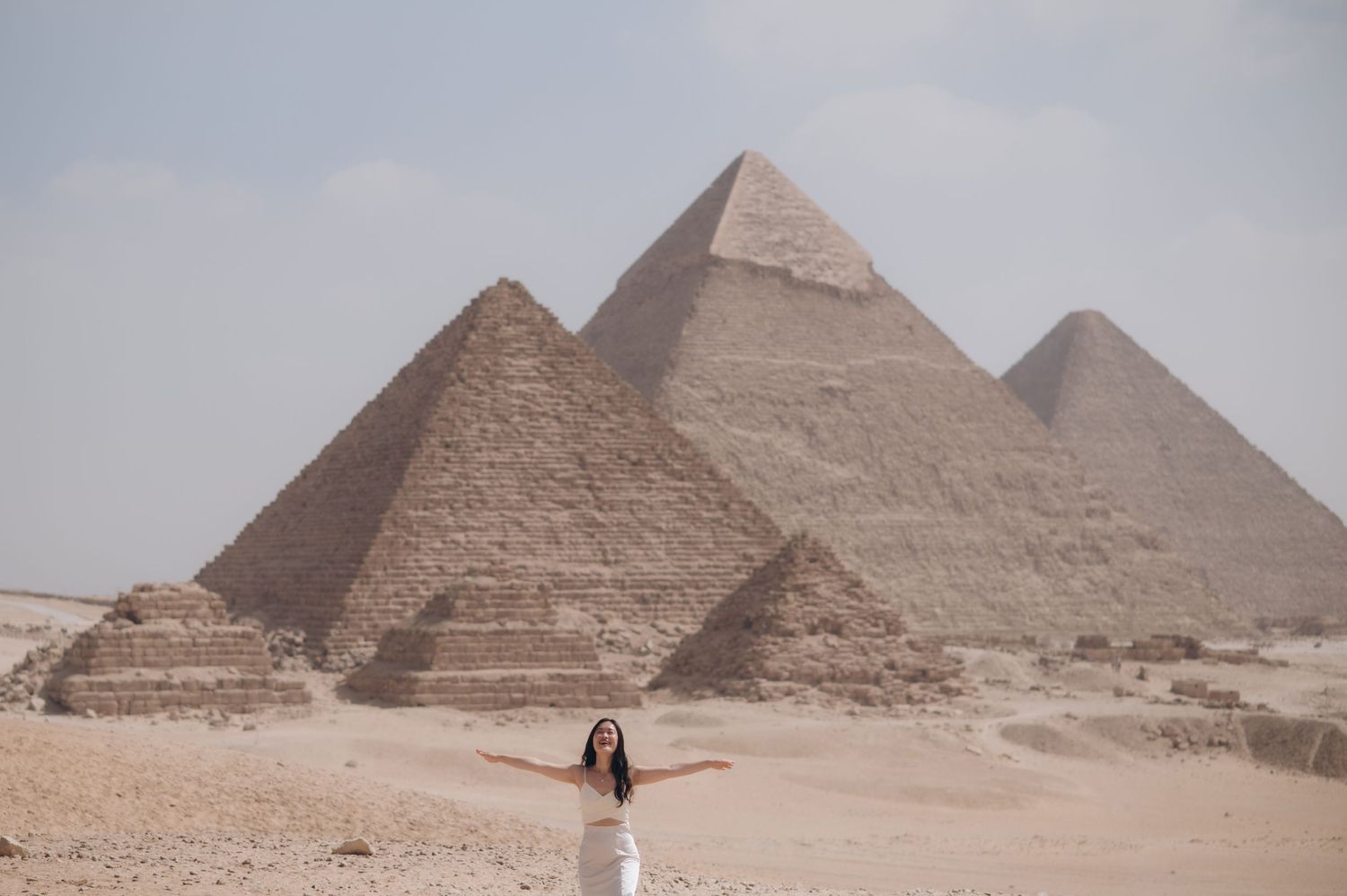 Tourist posing with arms outstretched in front of the three Great Pyramids of Giza.