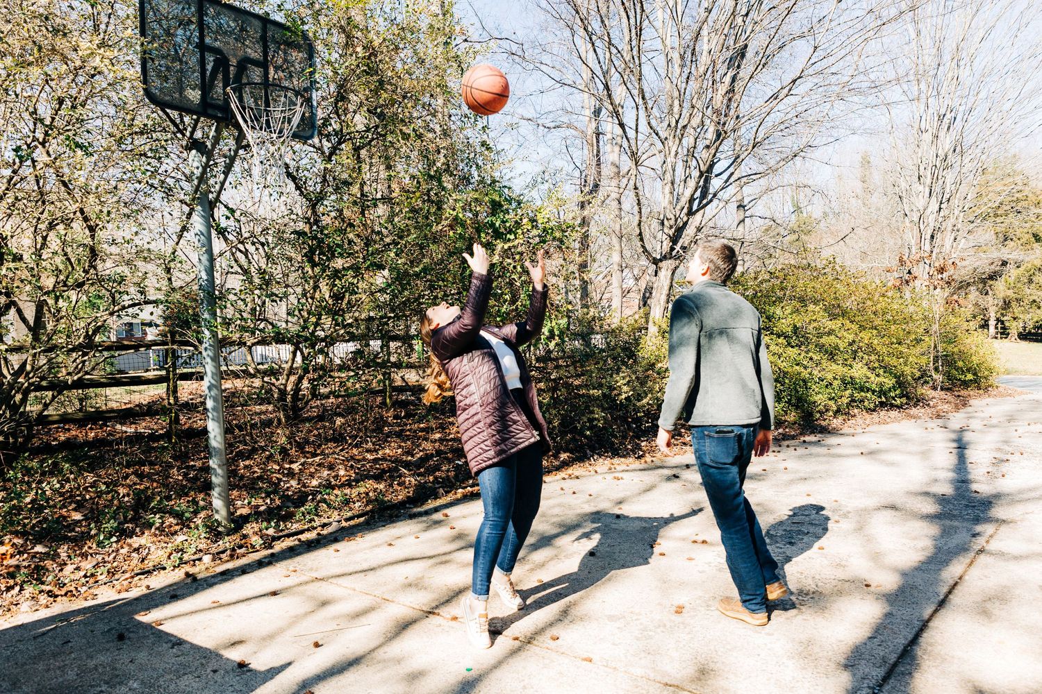 Two people walking side by side on a paved path surrounded by trees casting shadows in sunlight playing basketball.