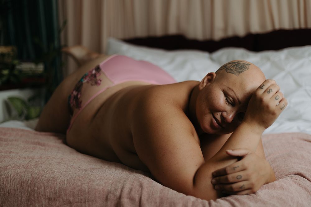 Artistic boudoir photo of a person wearing pink lingerie resting sensually on white bedding in natural light.