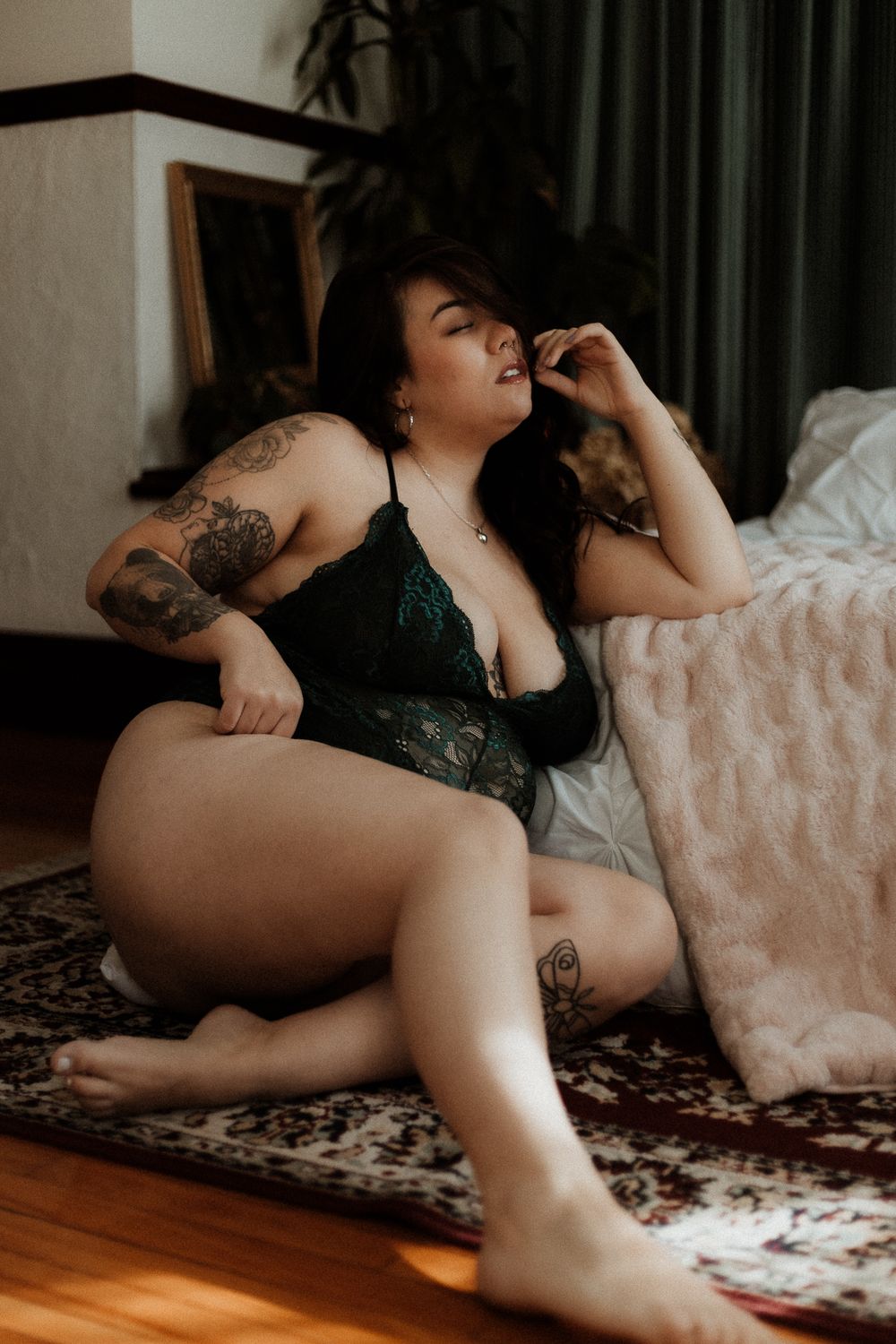 Artistic boudoir photo of person in green lace lingerie reclining on patterned rug in moody lighting.