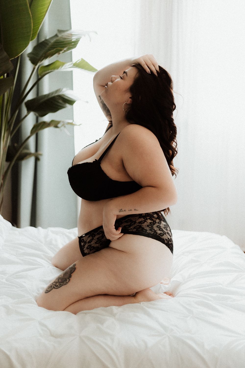 Curvy figure wearing black lingerie poses elegantly on white bedsheets near houseplant and window.