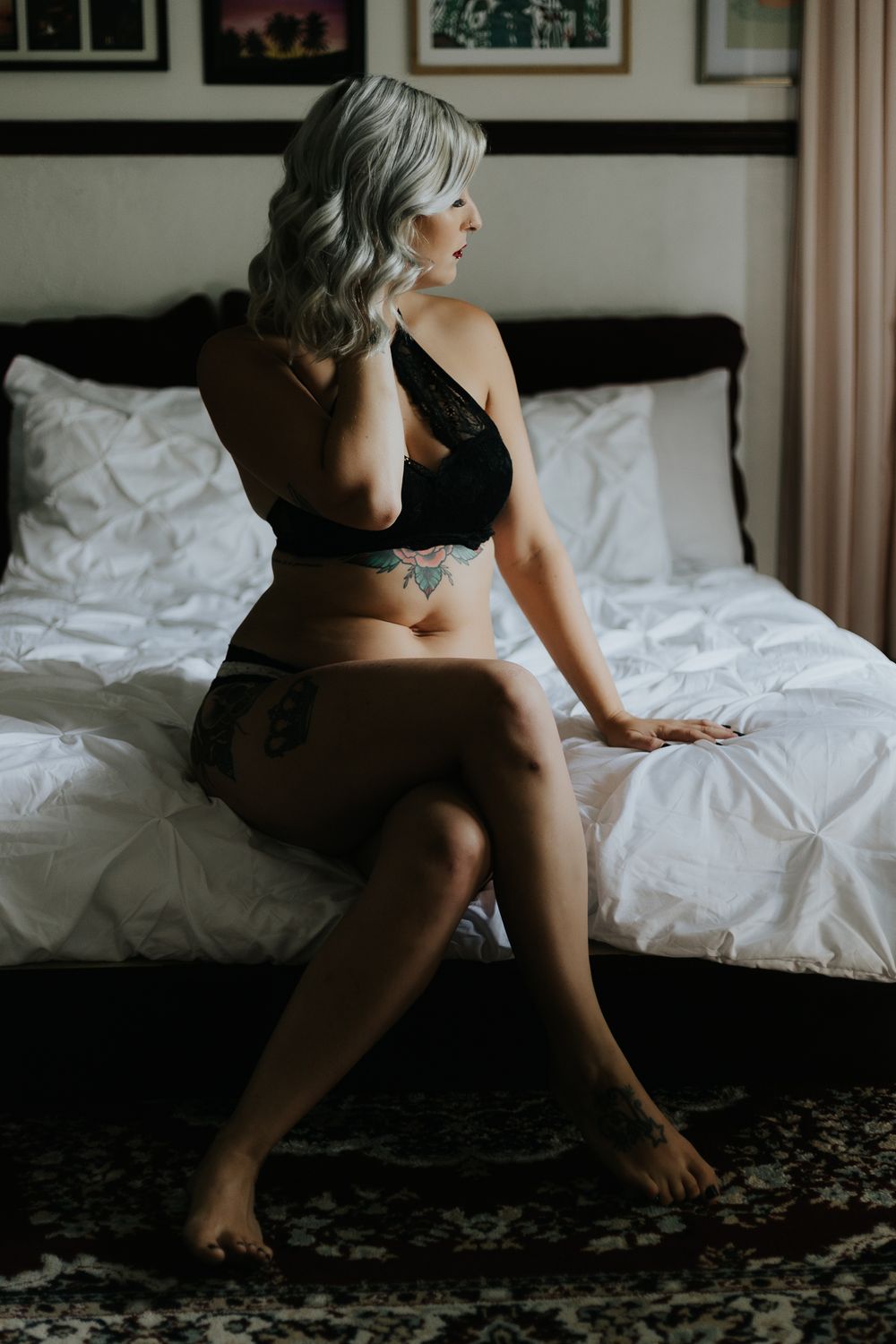 Intimate portrait of someone in black lingerie sitting on white bedding with silver hair and visible tattoos.