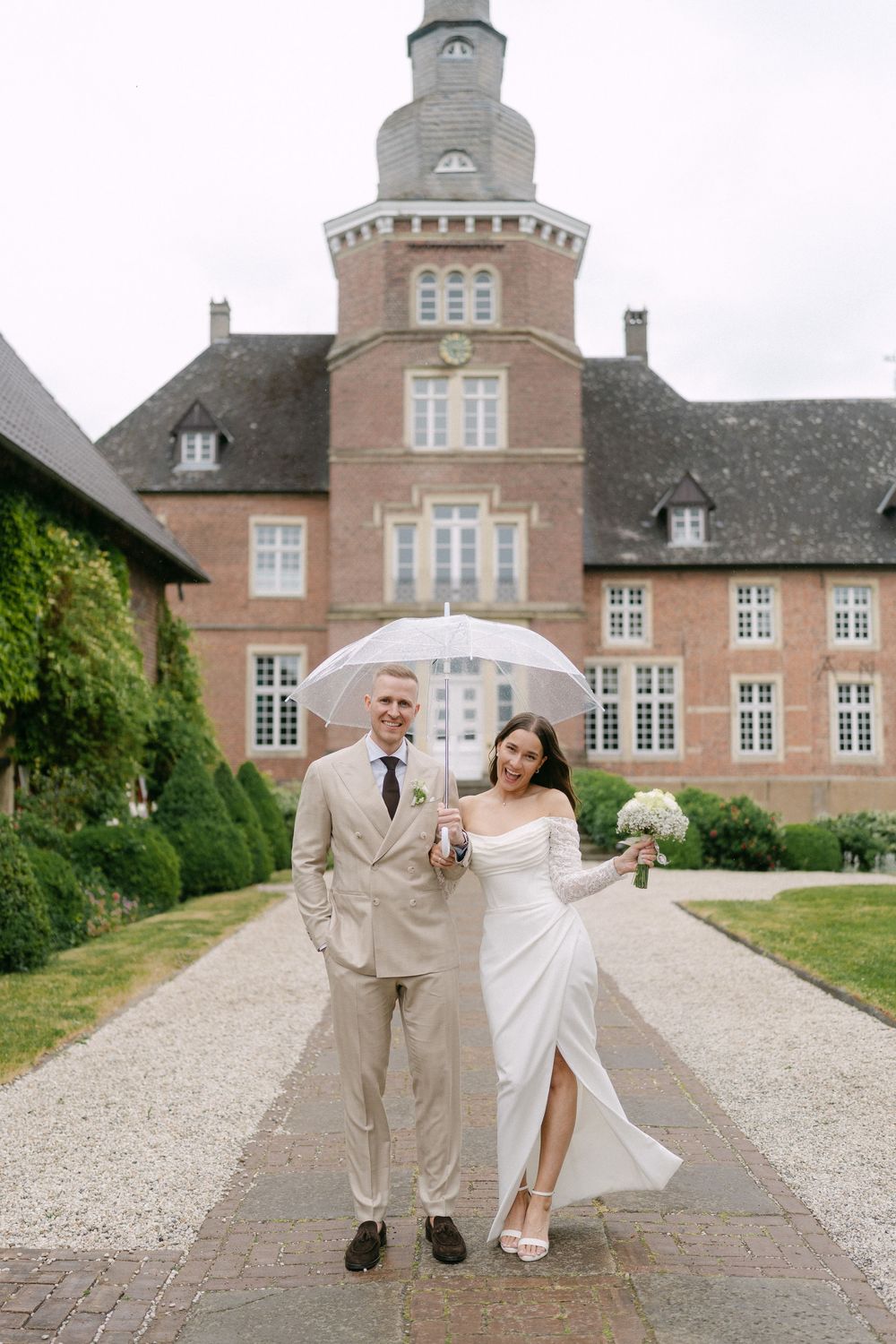 A wedding couple poses with an umbrella in front of a grand European-style mansion.