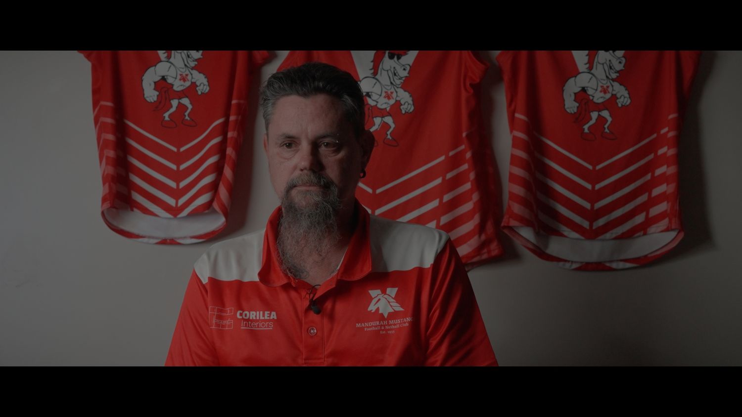 A person in a red polo shirt stands against a wall with red and white banners displaying heraldic symbols.