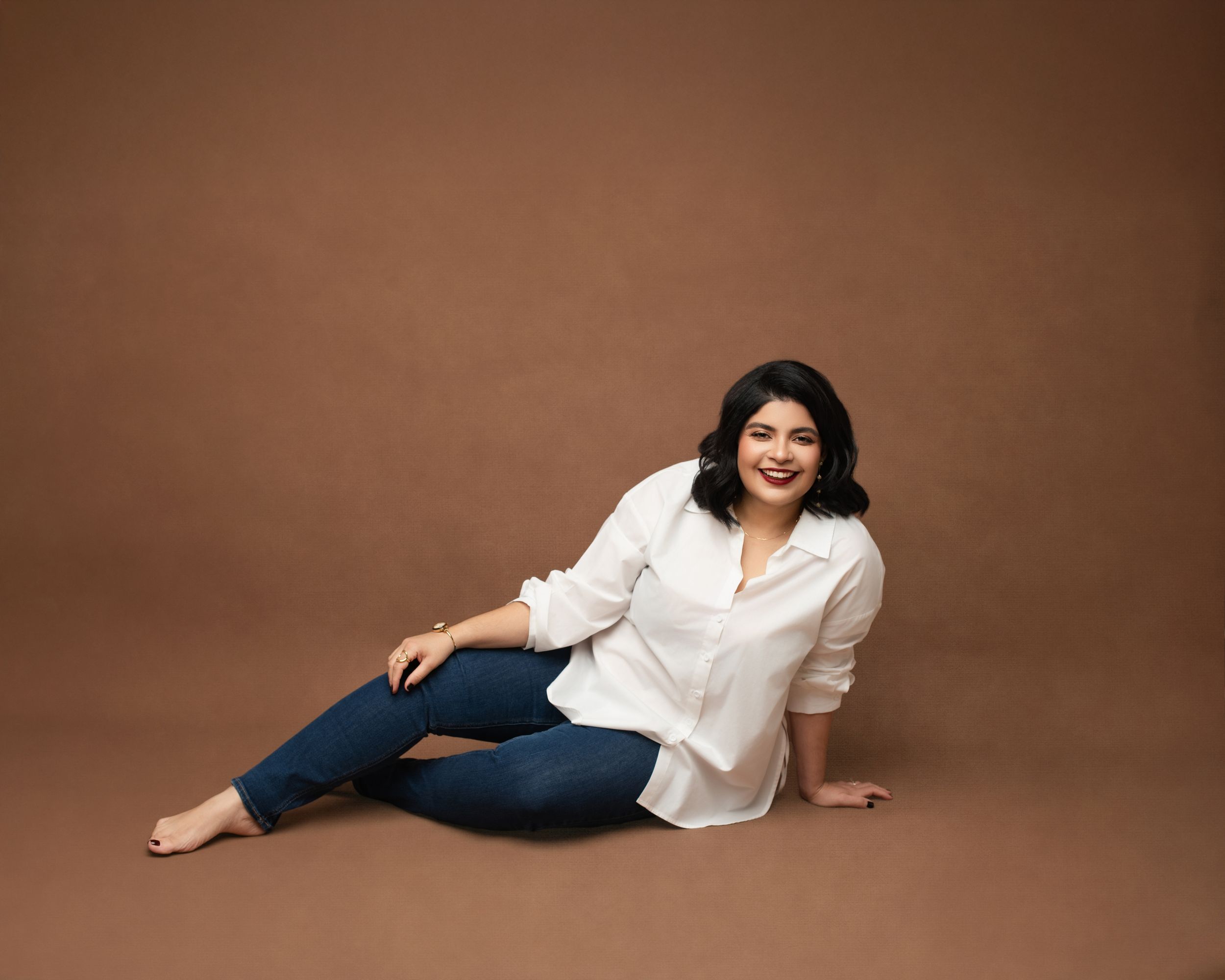 A person in a white button-down shirt and jeans poses casually while sitting on a brown backdrop.