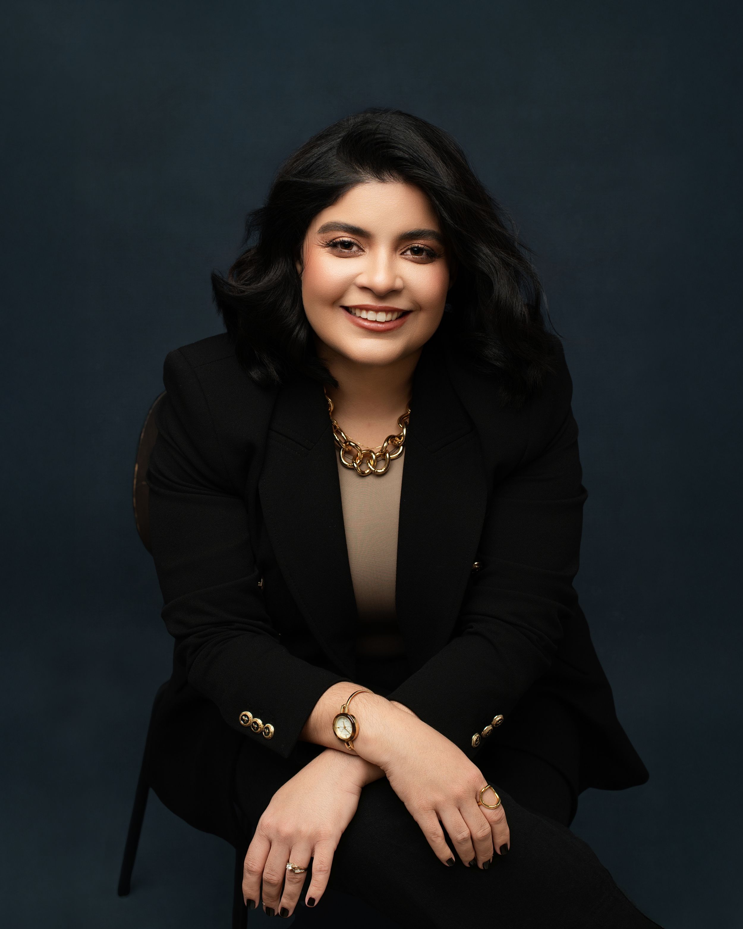 Professional studio portrait against dark background featuring someone in a black blazer with gold jewelry and warm smile.