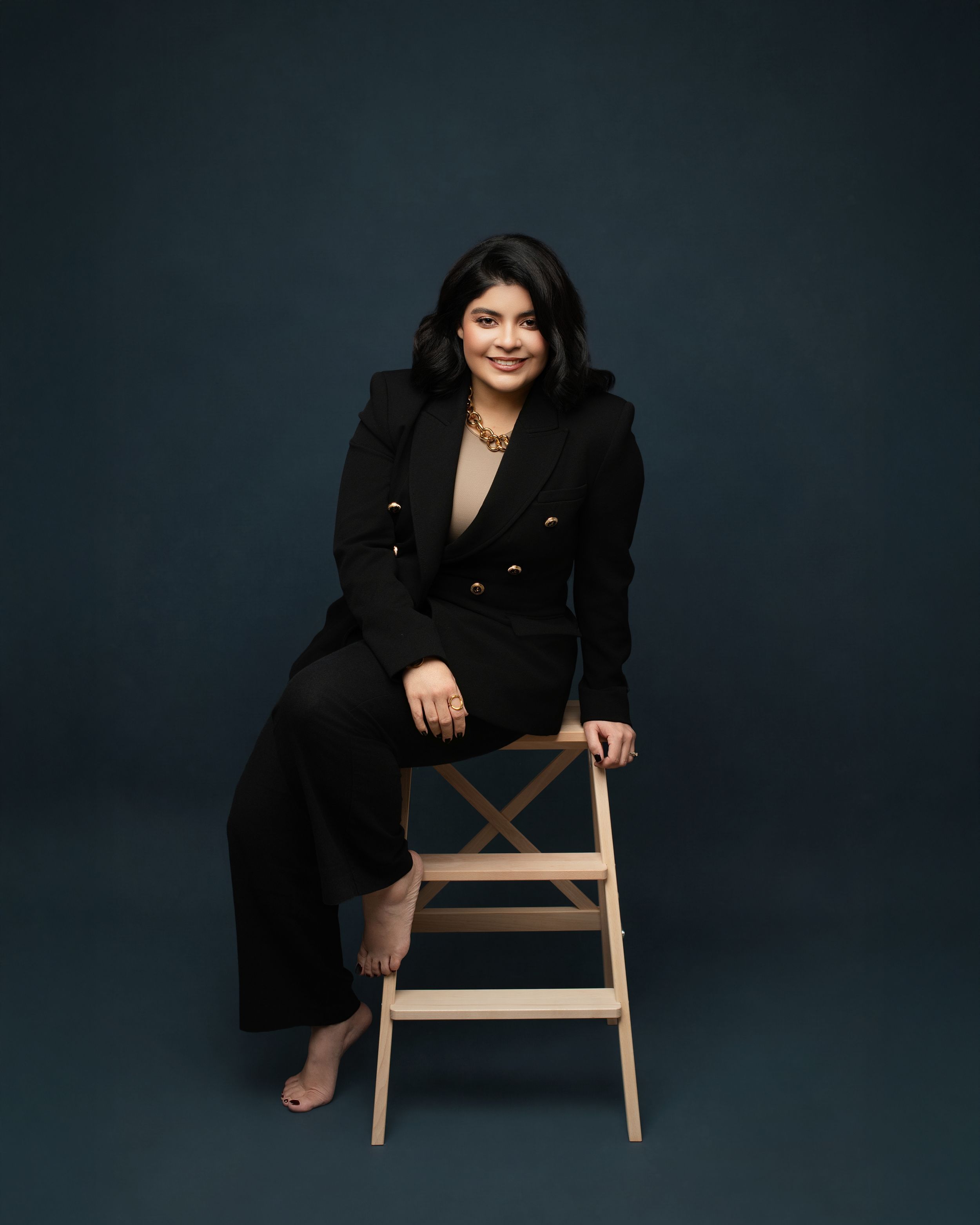 Professional studio portrait of someone in a black suit seated on a wooden chair against a dark backdrop.