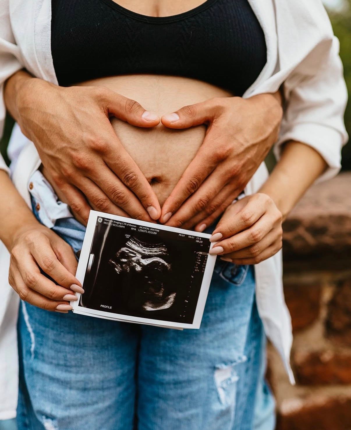 Person holds a sonogram photo while forming a heart shape with hands over their belly wearing jeans and black top.