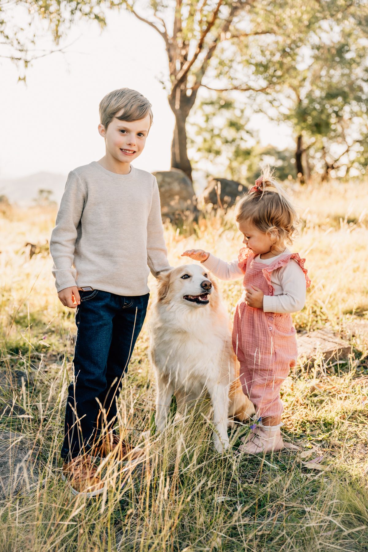 A young boy and his sister pat their dog outdoors in the golden sunlight.