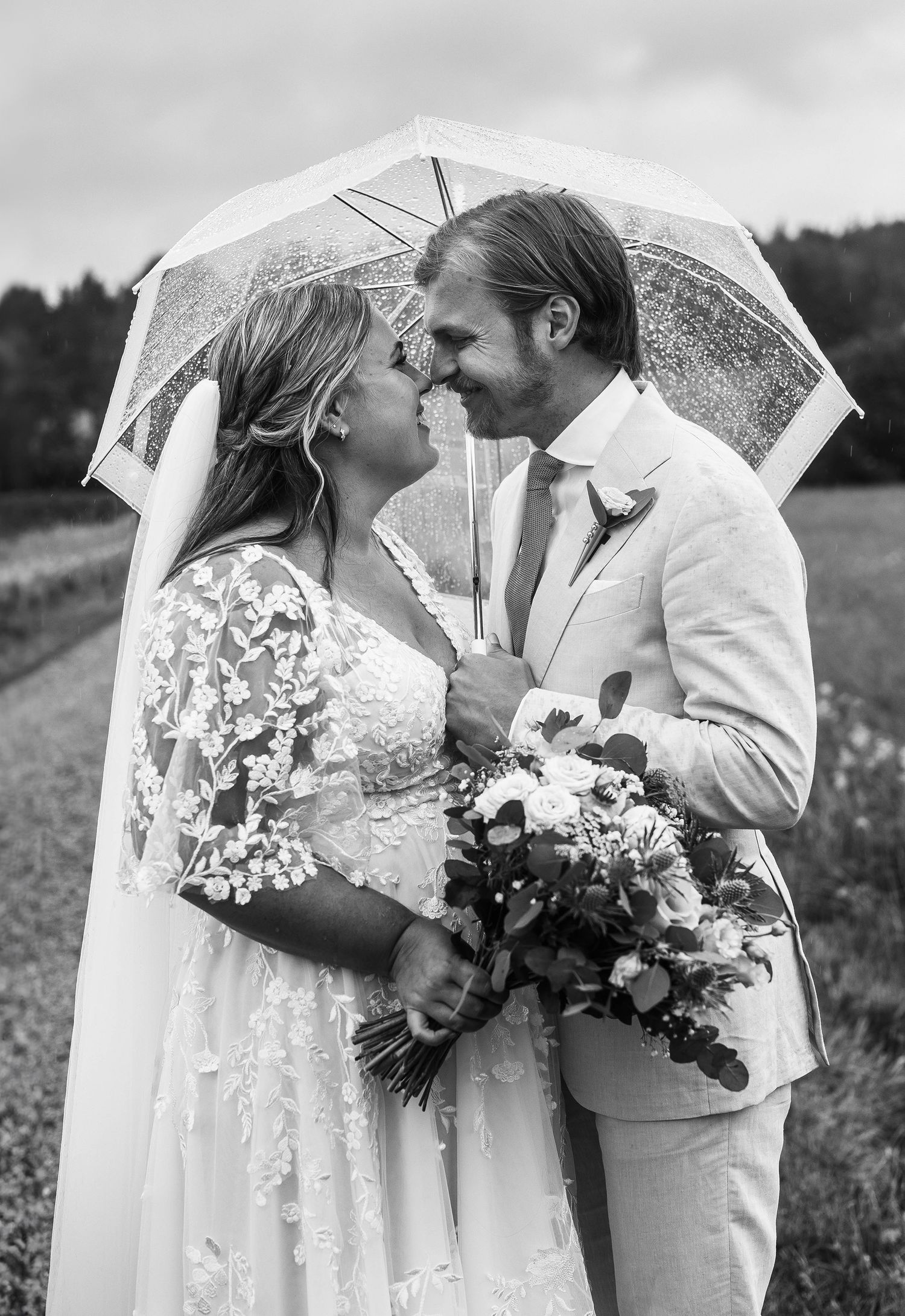 Svartvitt porträtt av ett gift par som står nära varandra i regnet under ett genomskinlig paraply i Trönö, Hälsingland