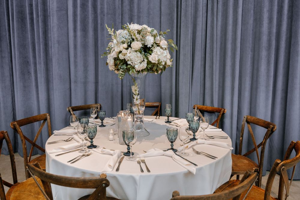 Elegant wedding table setting with white floral centerpiece, wooden crossback chairs, and blue drapery backdrop.