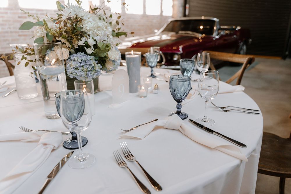 An elegant wedding reception table with blue glassware, white linens, and floral centerpieces in an industrial venue with vintage car.