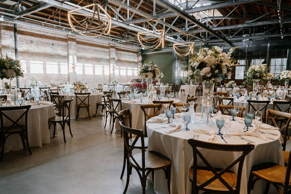 Elegant wedding reception venue with round tables, crossback chairs, and tall floral centerpieces in an industrial space.