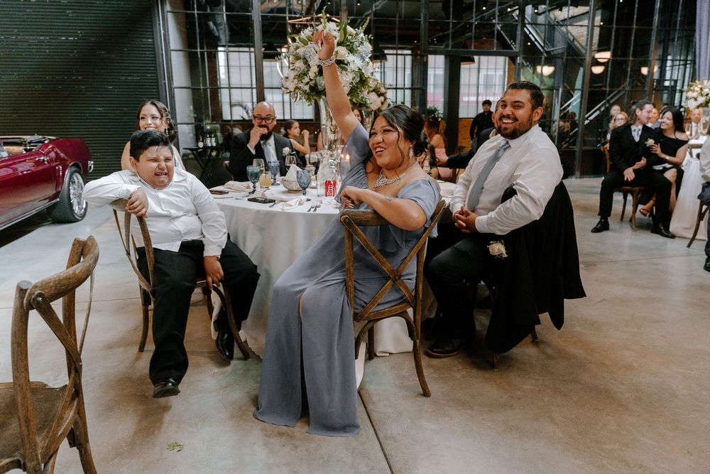 Wedding reception guests celebrate joyfully at a modern industrial venue with round tables and wooden cross-back chairs.