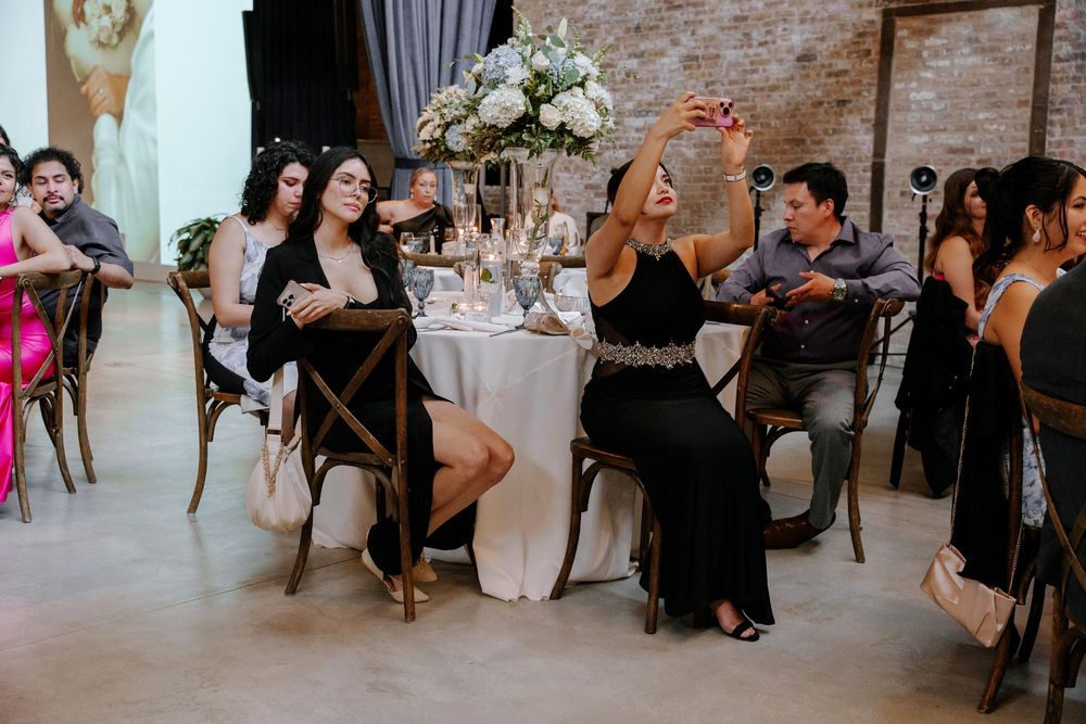 Wedding guests socialize at a rustic brick-walled reception venue with elegant decor and dark curtains.