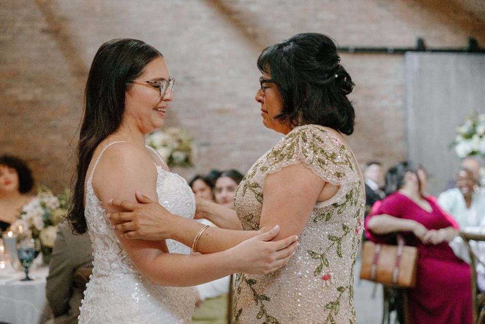 Two people wearing floral and white dresses embrace emotionally during a wedding ceremony in a brick-walled venue.