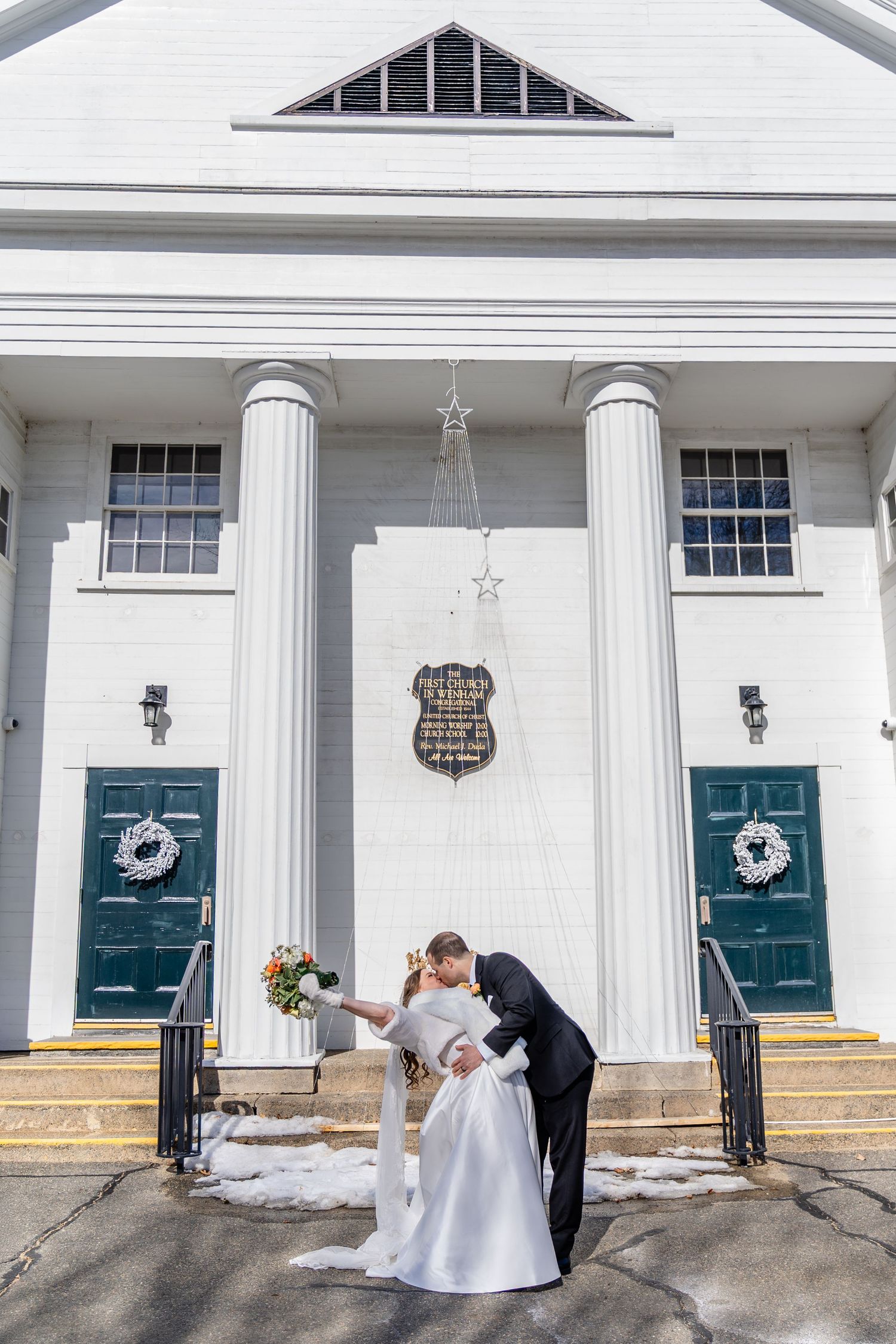 Wedding couple poses in front of white church building with twin green doors and classic columns in winter.