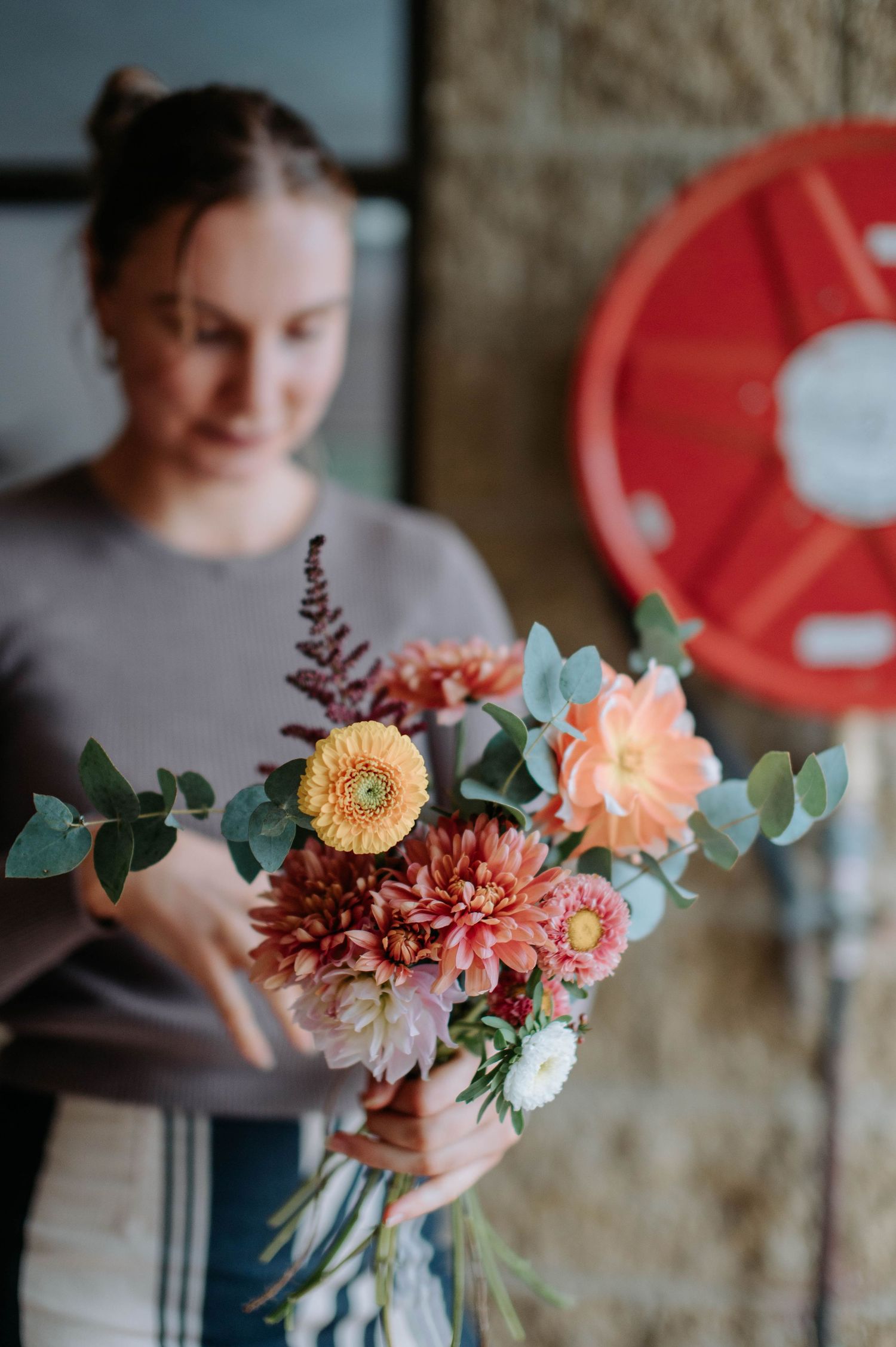 Florist arranging coral and peach colored dahlias with eucalyptus in vase against brick wall background.
