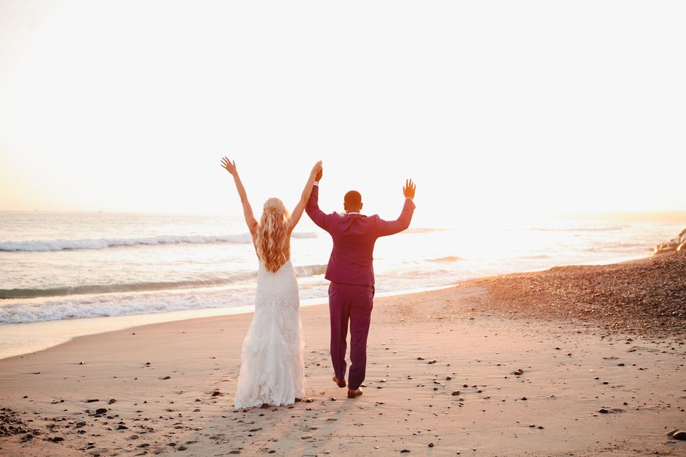 A couple in wedding attire walks along a beach at sunset with waves crashing in the background.