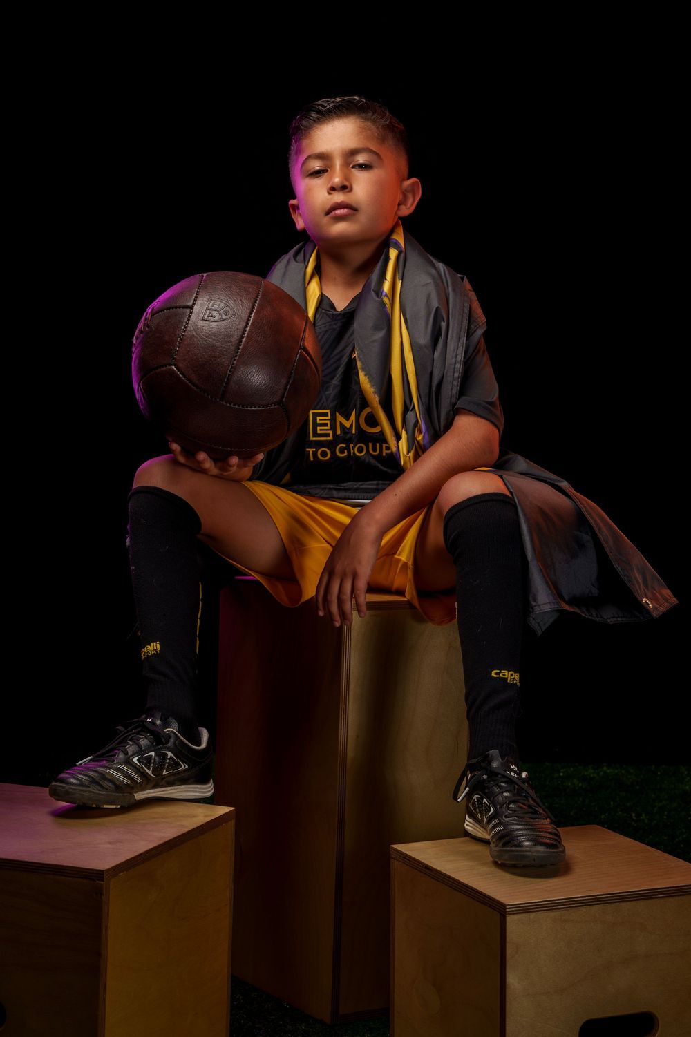 Youth athlete with soccer ball sits on brown cube against dark backdrop.