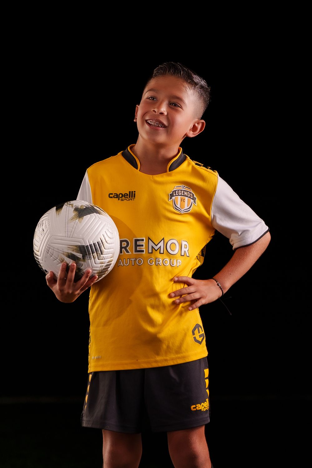 Young athlete poses with soccer ball wearing yellow and black Remor team uniform against dark background.