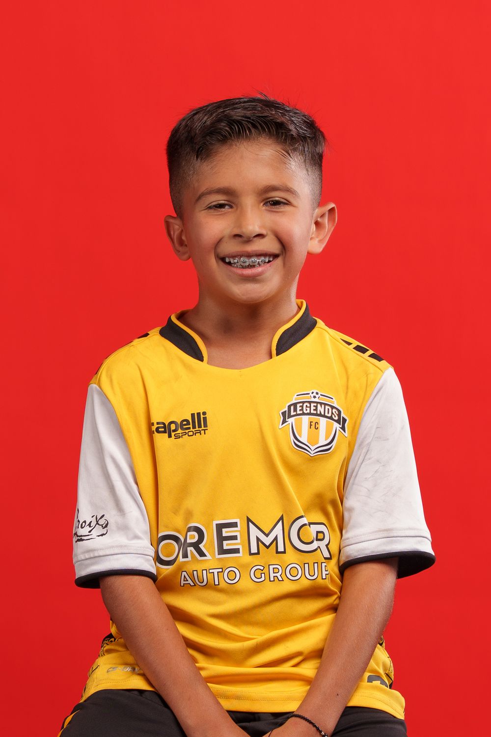 Young soccer player in yellow Legends FC team jersey poses playfully against red background.