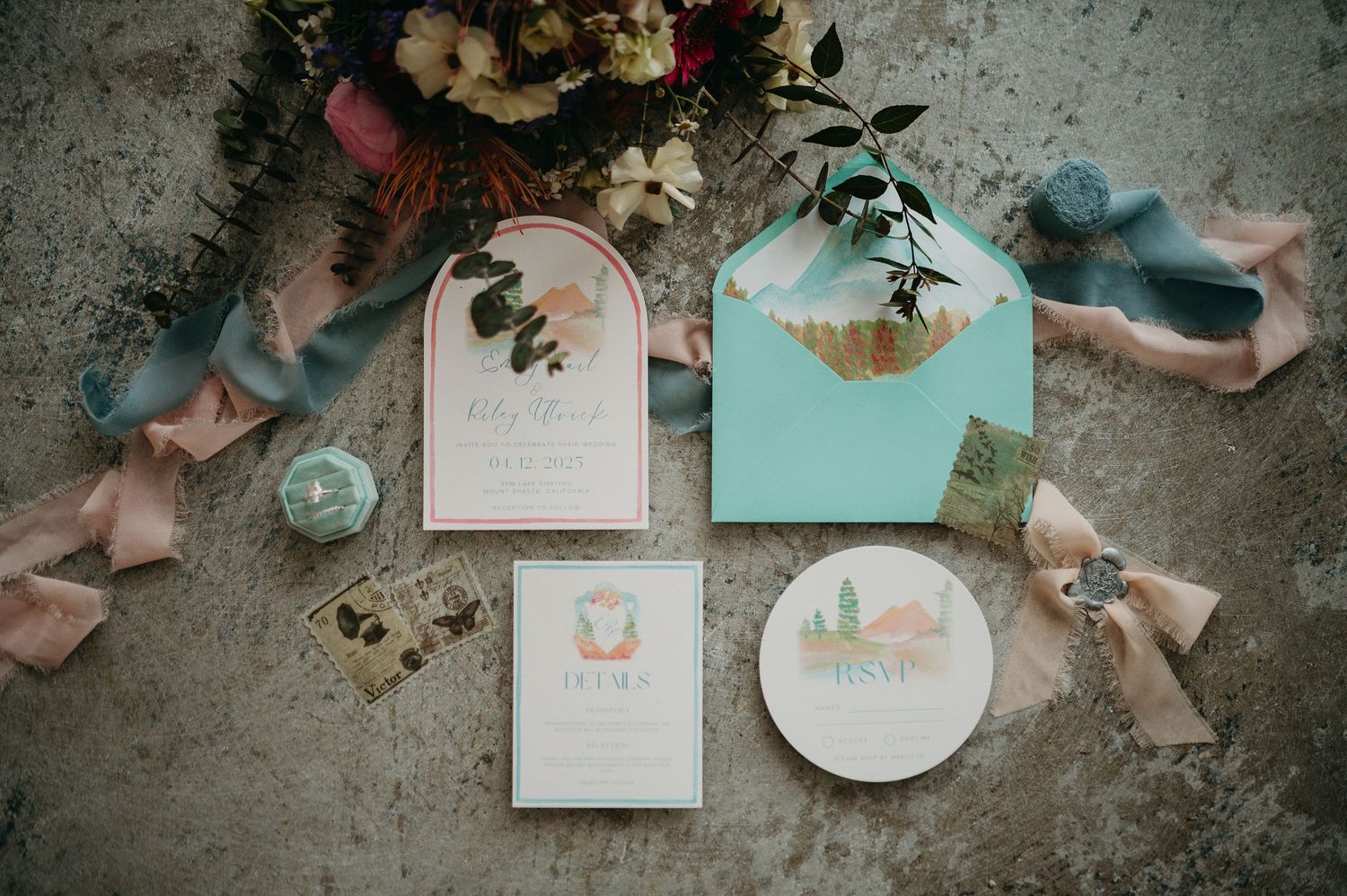Elegant wedding invitation suite with blush pink and mint green envelopes, ribbons, and floral decorations on concrete surface.