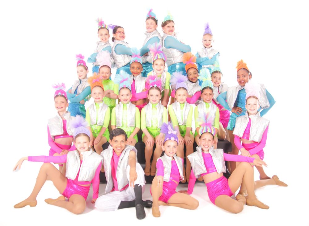 Colorful dance group performs synchronized jumping poses in bright costumes.
