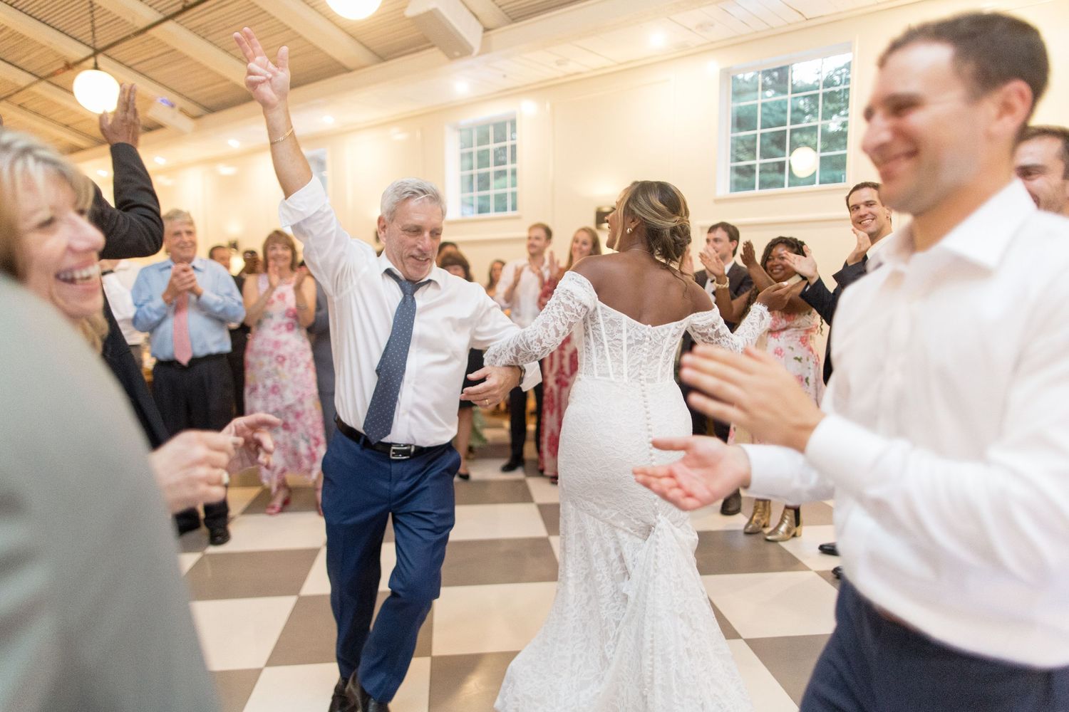 Wedding guests dance together on a wooden floor beneath exposed ceiling beams at an indoor venue.