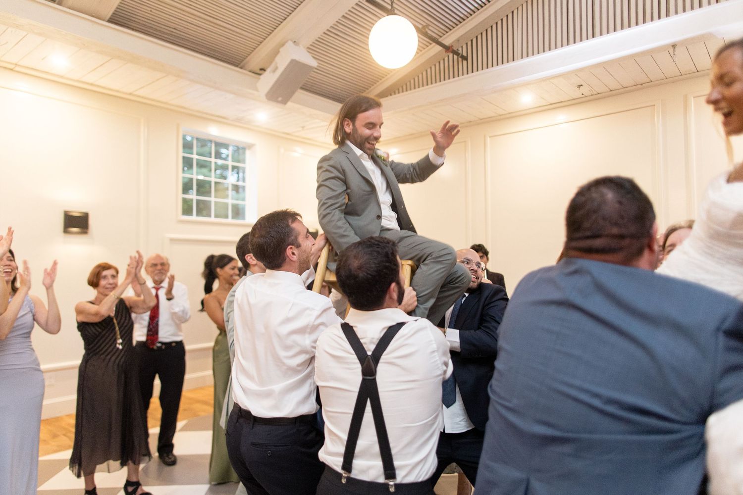Party guests dancing together with raised arms in a bright white reception hall with wood beam ceiling.
