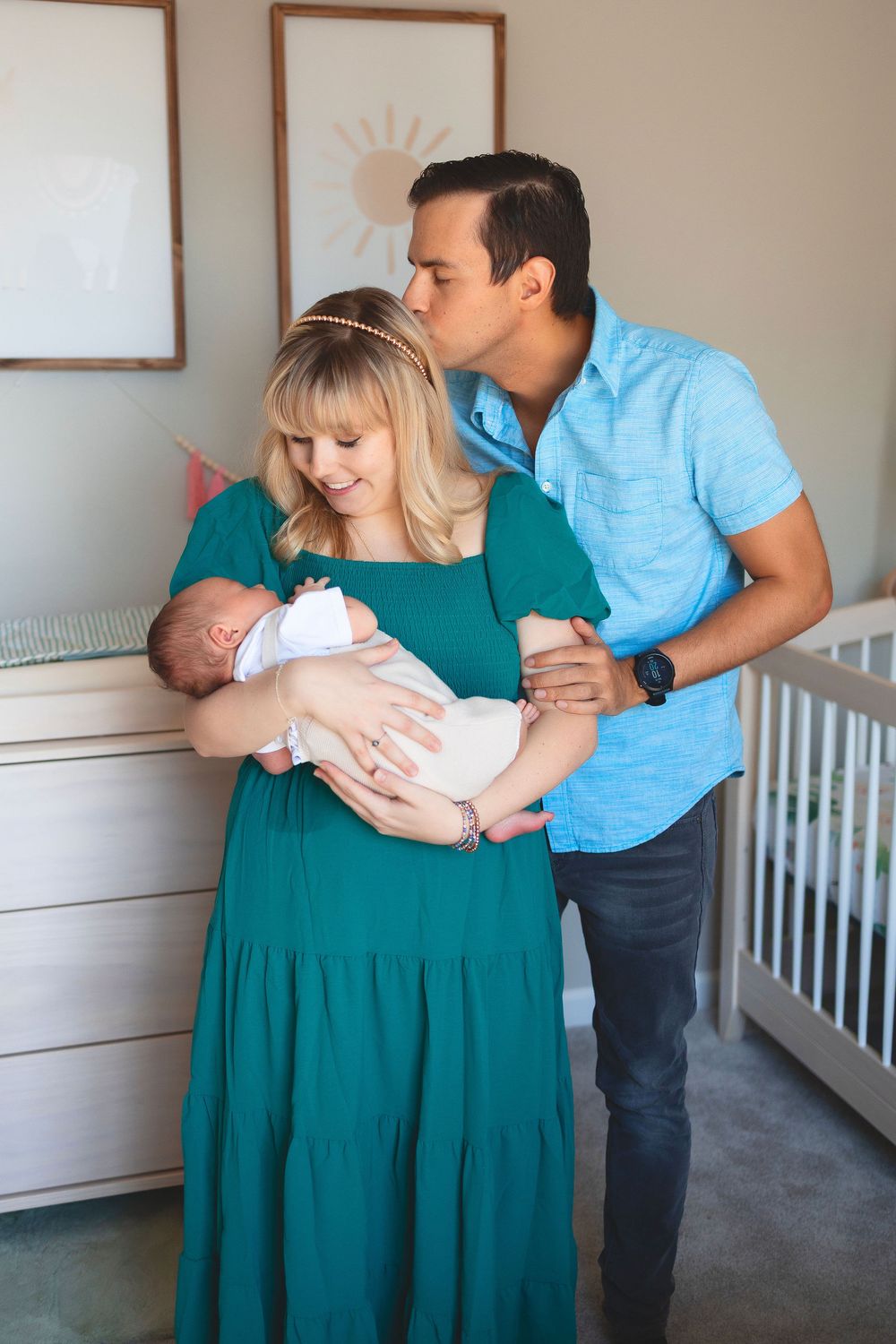 A loving couple shares a tender moment in a nursery while holding their newborn baby in a green dress and blue shirt.