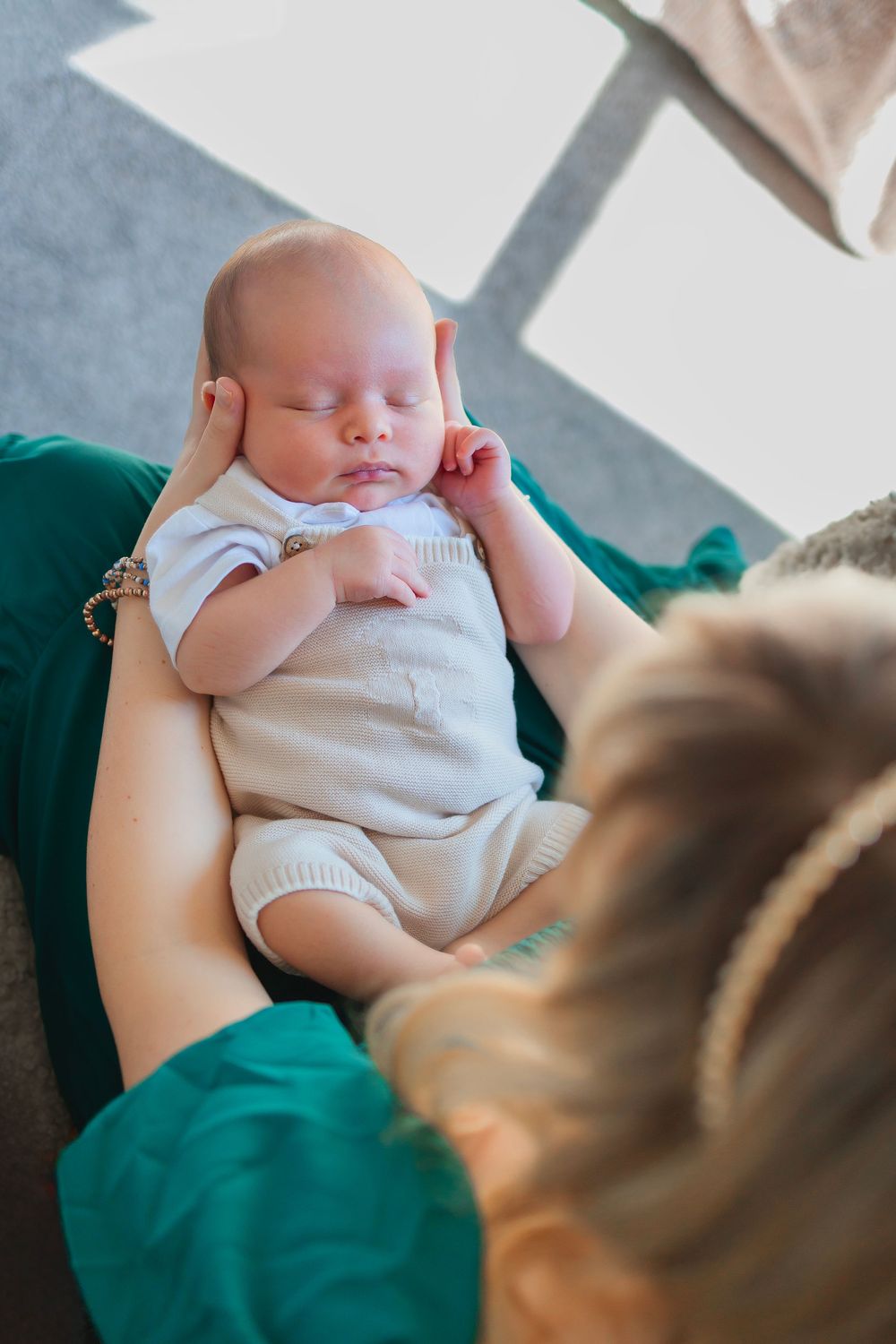 A sleeping newborn baby in white clothing rests peacefully on mothers lap.  Mother holding him in her forearms, she is looking down at him.