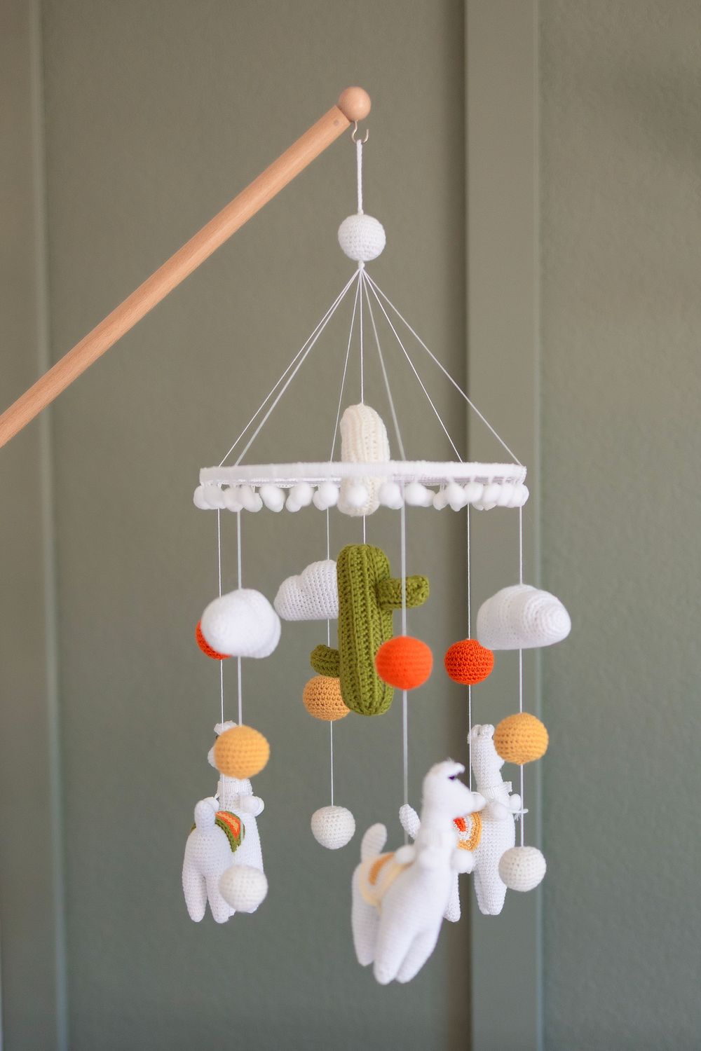 A baby mobile with white felt ghosts, orange and green cactus decorations, and pom poms hanging from a wooden dowel.