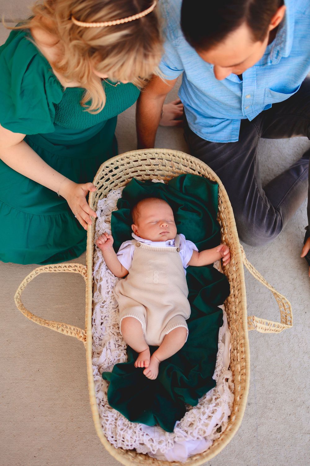 A sleeping baby lies in a woven moses basket with emerald green blanket while parents look down lovingly.