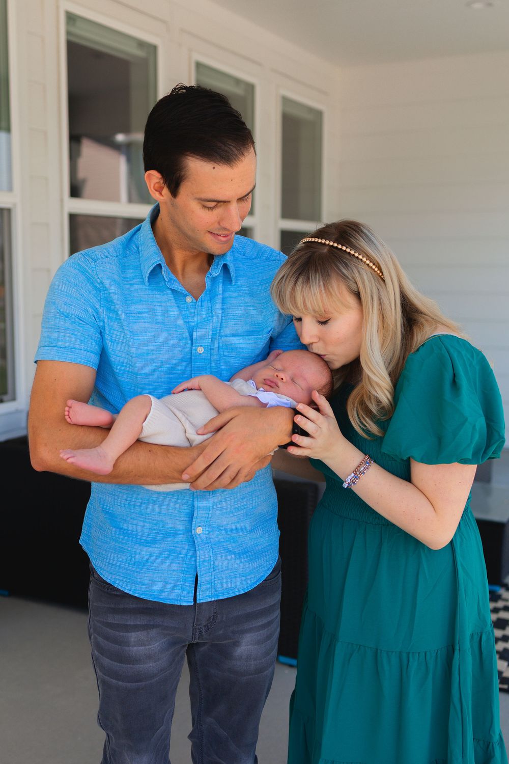 A couple in a blue shirt and teal dress share tender moments while holding their newborn baby.