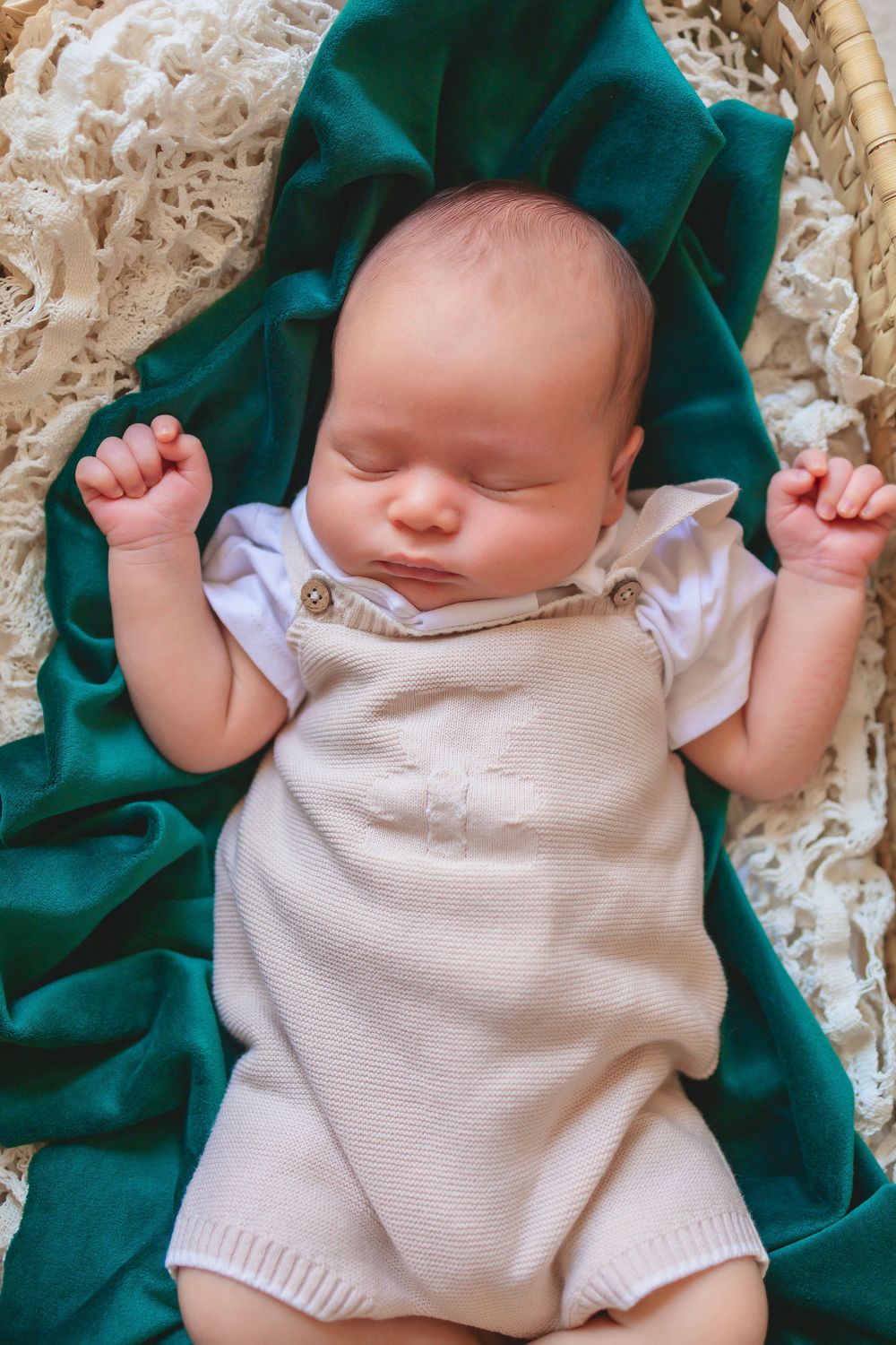 A sleeping newborn baby wearing a beige knit romper rests peacefully on a textured white blanket with teal fabric accents.