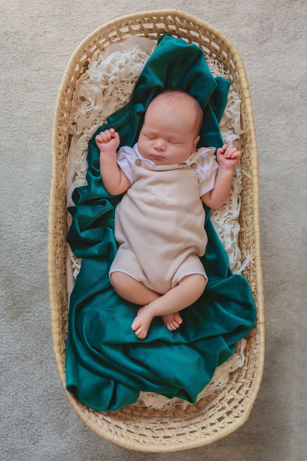 Sweet newborn rests peacefully in a woven basket lined with emerald green fabric while wearing a soft beige onesie.