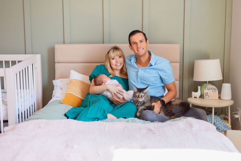 A couple holding a newborn relaxes on a bed with their cat in a cozy bedroom setting with a white crib in the background.