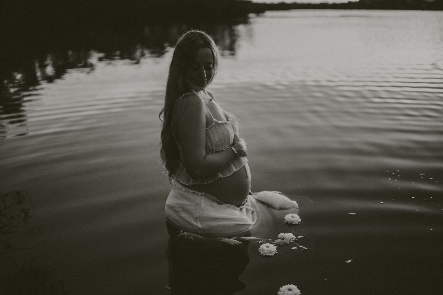 A person sits peacefully by a tranquil lake at sunset, creating ripples in the dark reflective water. sarasota maternity photographer