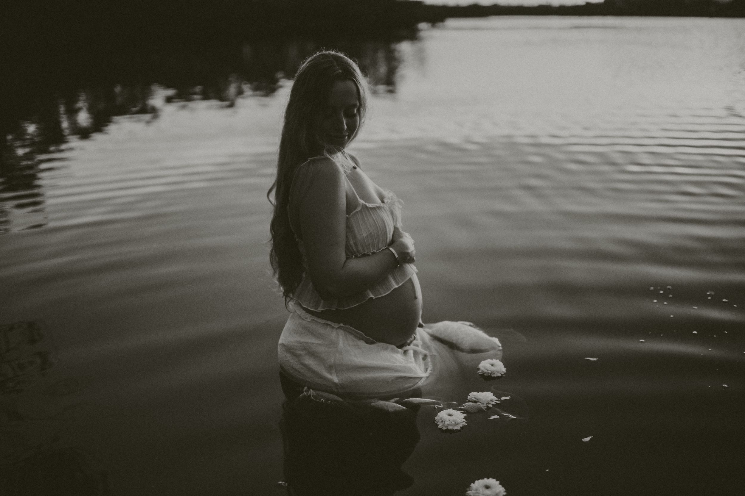 A person sits peacefully by a tranquil lake at sunset, creating ripples in the dark reflective water. sarasota maternity photographer