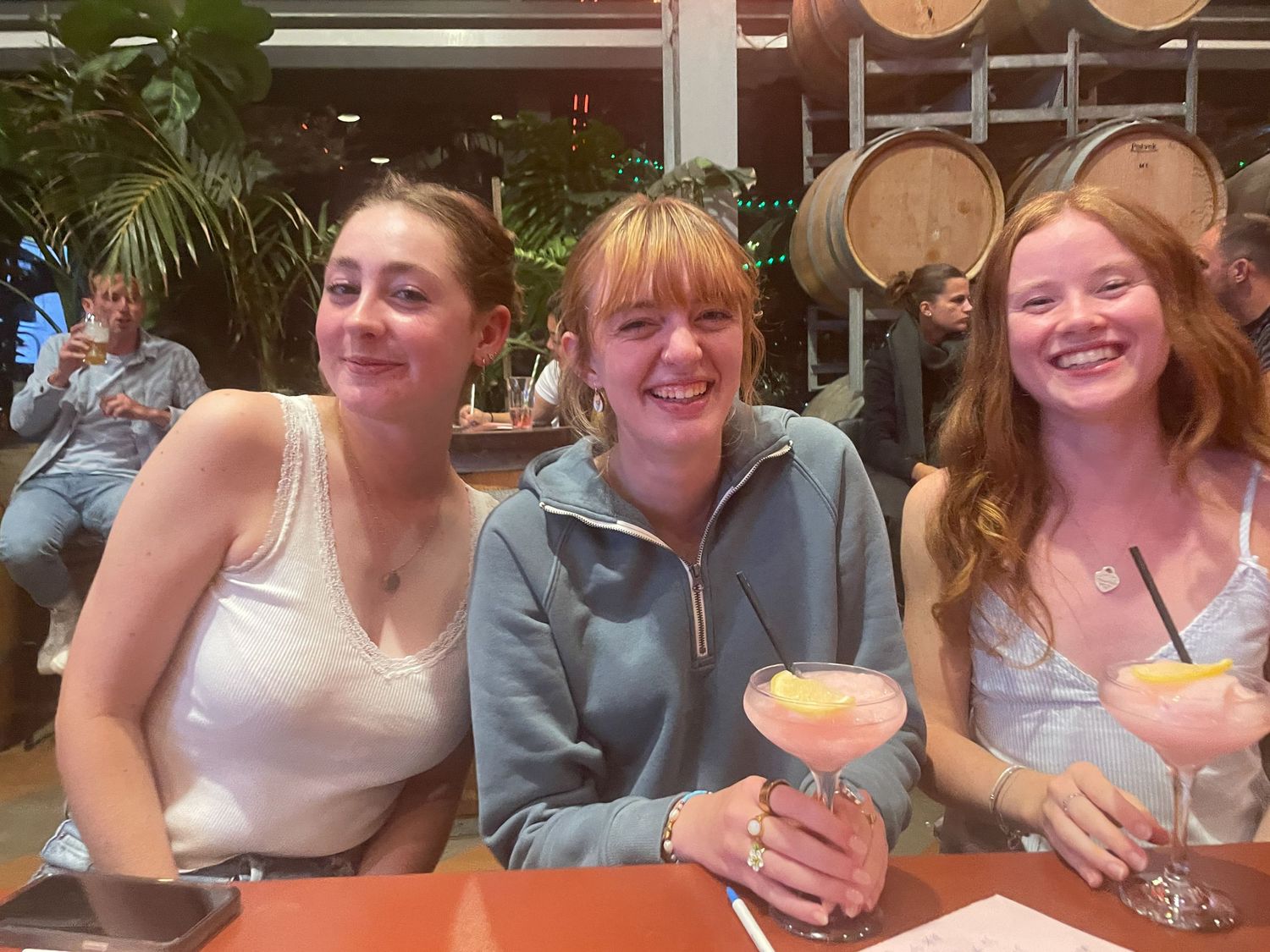 Three friends enjoying cocktails together while seated at a restaurant with wooden wine barrels in the background.