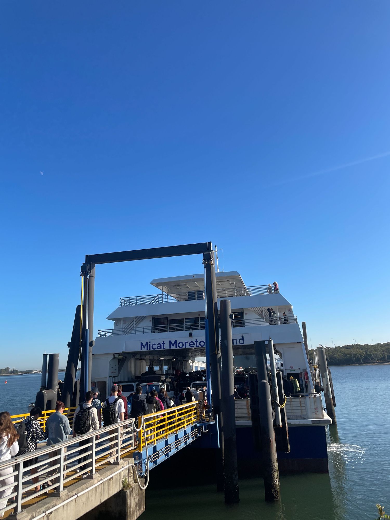 Ferry passengers board a vessel at a dock under bright blue skies with water and docking structures visible.