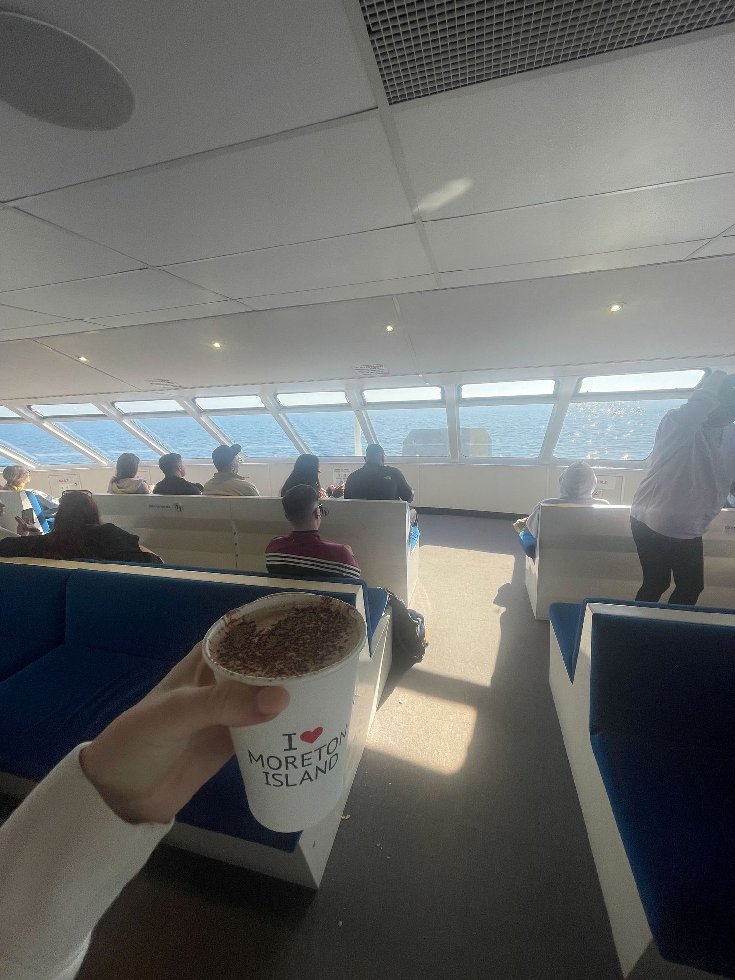 Passenger holds coffee cup inside ferry cabin with blue sky and ocean views through slanted windows during daytime sailing.