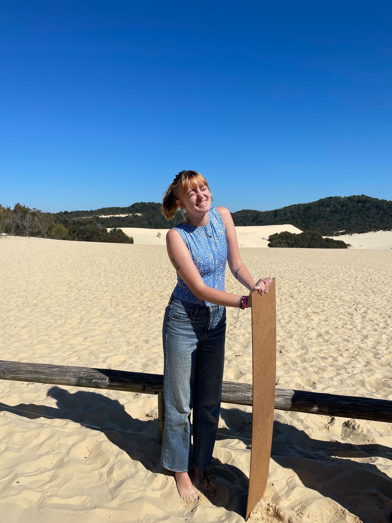 Someone in a blue sleeveless top and jeans stands against a wooden post on sand dunes under bright blue sky.