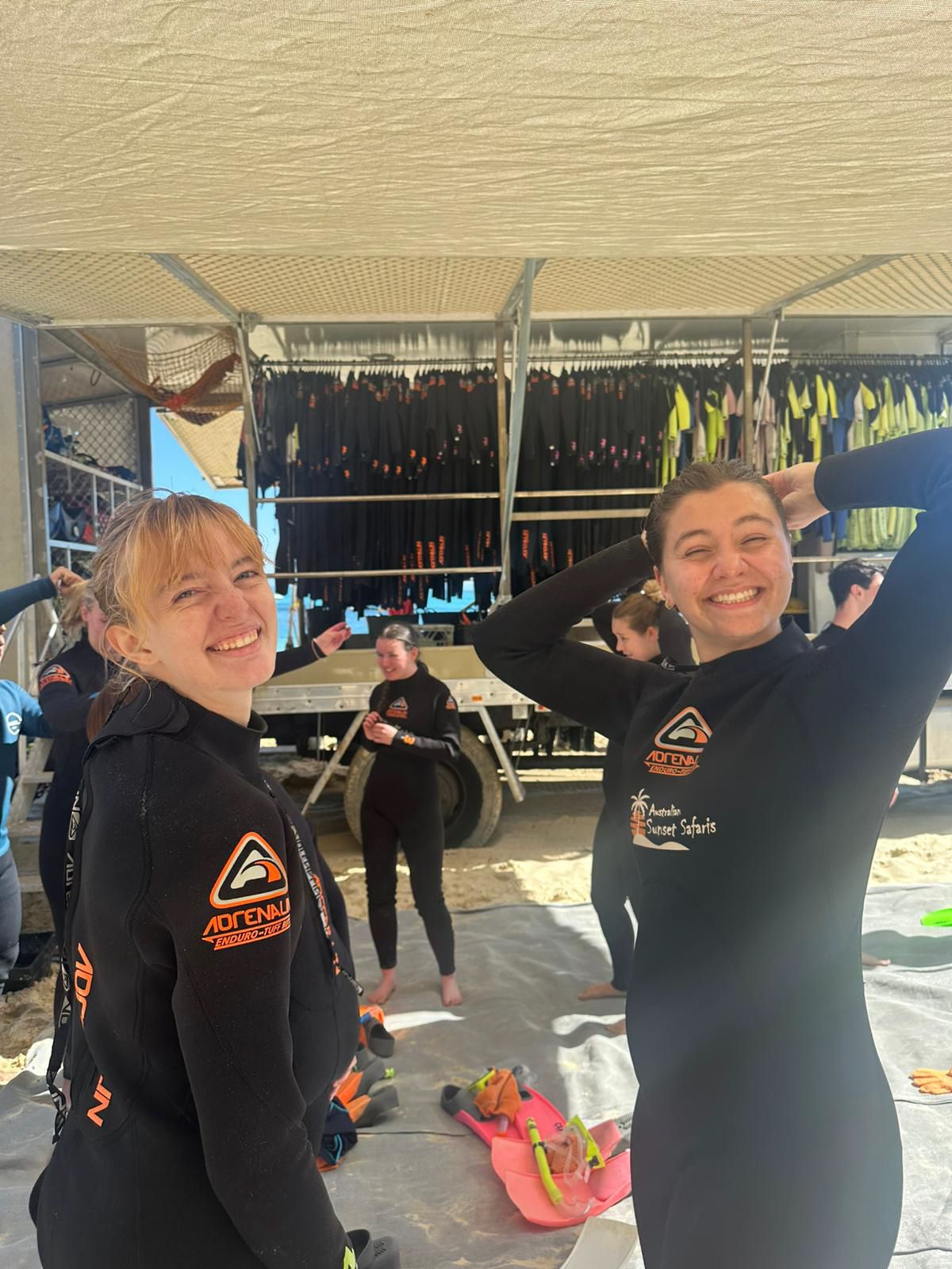 Two people in black wetsuits smiling and posing together under a beachside canopy with surf gear visible in background.