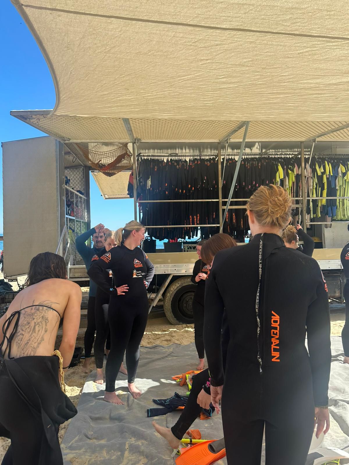 A group in wetsuits gathers on the beach by a truck preparing for scuba diving or water activities.