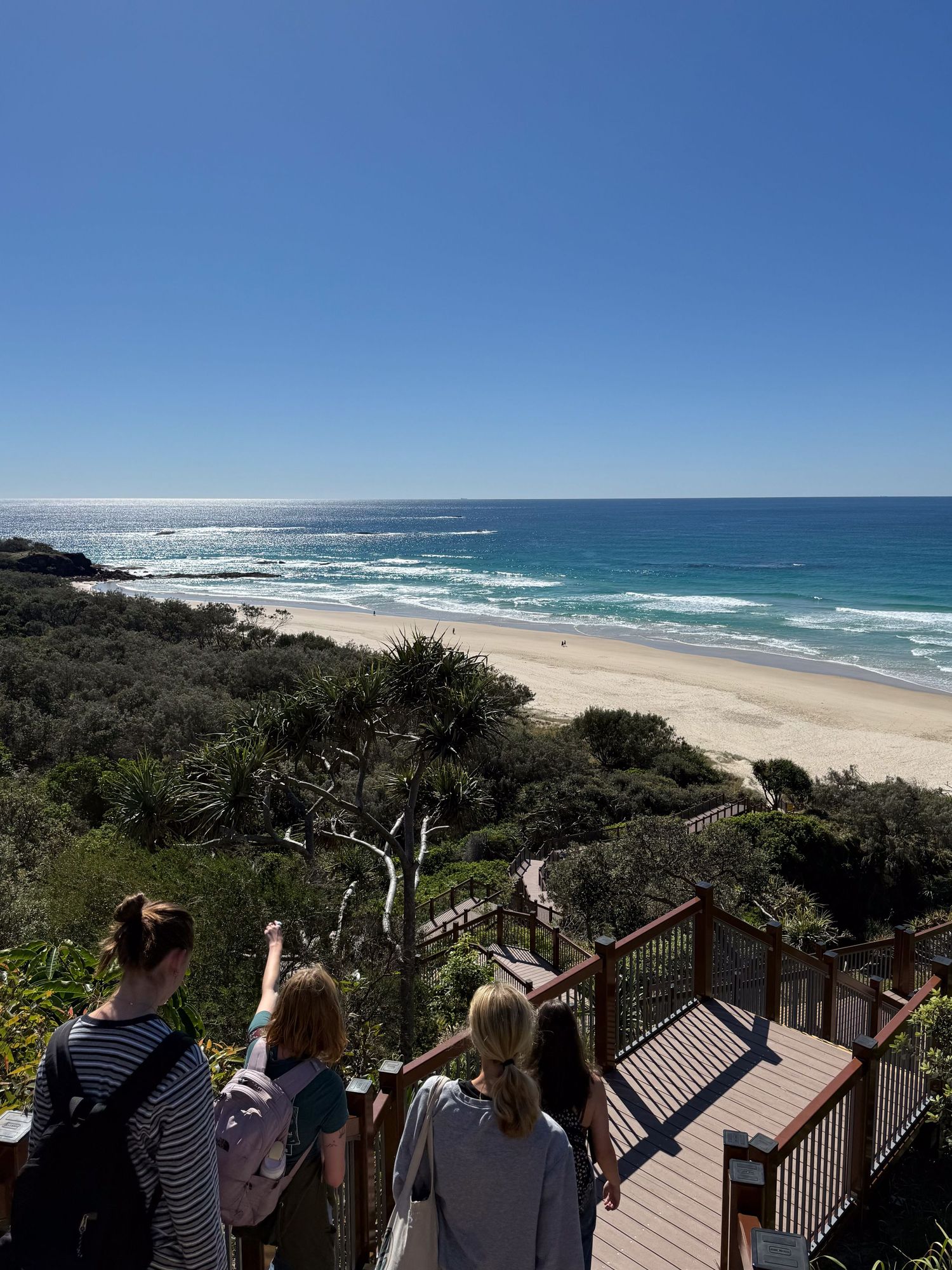 Tourists on an observation deck overlooking a pristine beach with turquoise waters and rocky coastline on a sunny day.