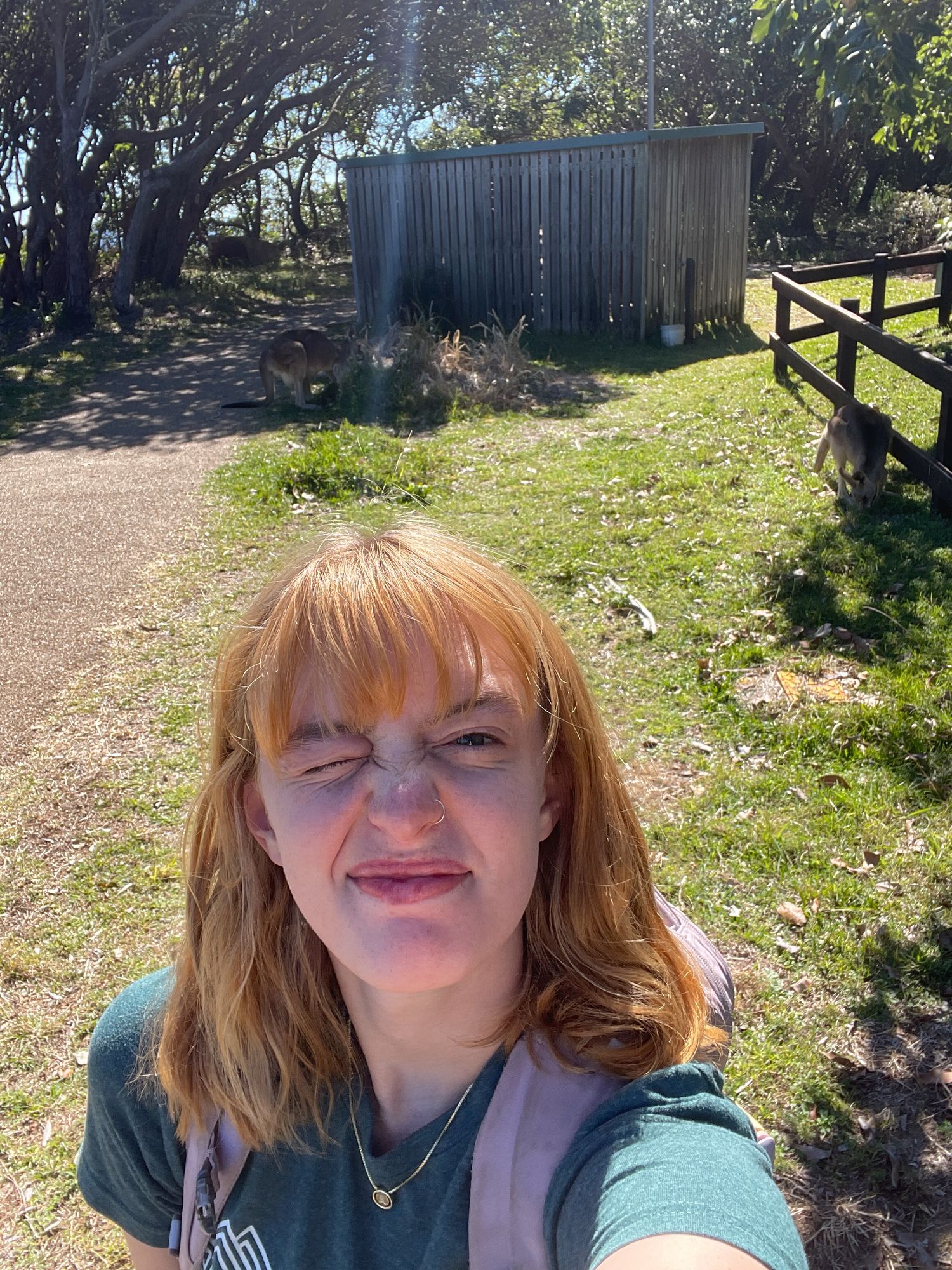 A sunny selfie taken outdoors with a metal storage shed and wooden fence visible in the garden background.